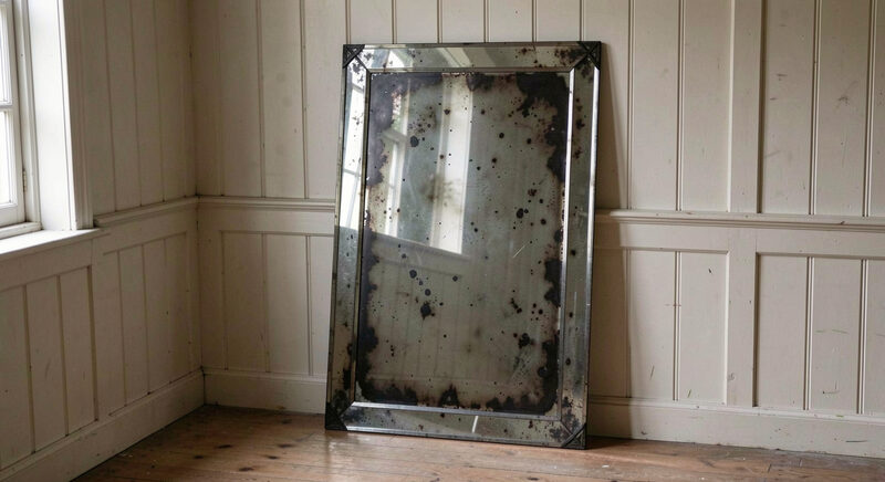 An old mirror with a bevelled glass border and darkened, spotted silvering, propped against a panelled wall in a quiet room, soft light from a side window.