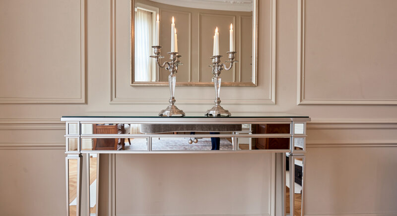 A mirrored console table against a panelled wall with a pair of candlesticks on top, its reflection doubling the room behind.